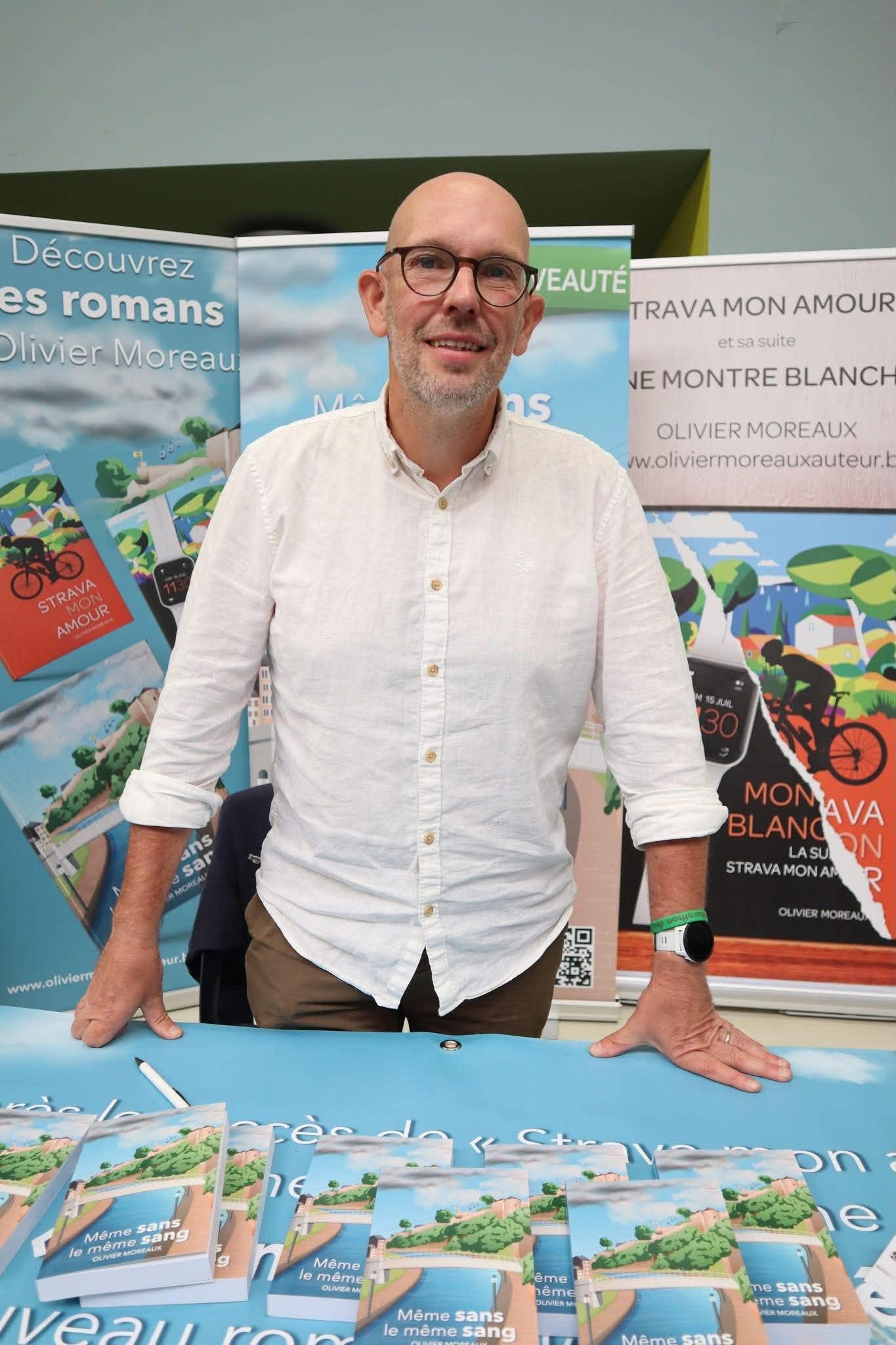 Olivier Moreaux, wit hemd, staand achter zijn signeertafel, omringd door zijn affiches en exemplaren van « Même sans le même sang »